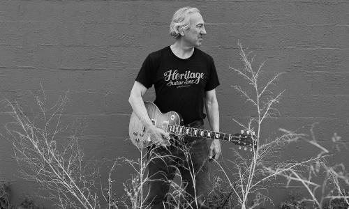 John Hern holding guitar in front of orange brick wall